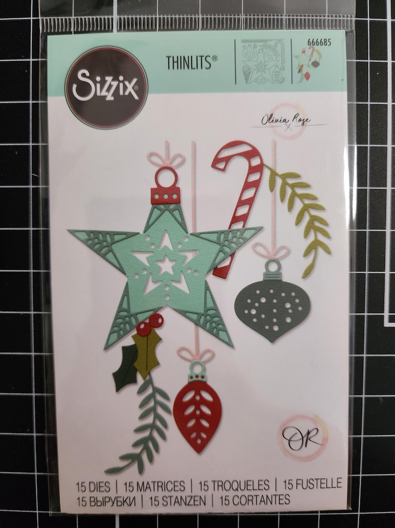 Puede incluir: Set de 15 troqueles Sizzix Thinlits 666685 de Olivia Rose. El empaque muestra formas troqueladas que incluyen un adorno de estrella verde claro, un bast&oacute;n de caramelo rojo y blanco, una bola gris, bayas rojas y follaje verde.