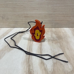 May include: A wooden pendant shaped like a flame with a yellow number "0" inside. The pendant is attached to a black cord.