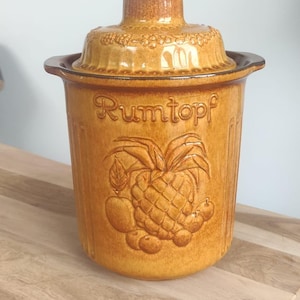 May include: A brown ceramic jar with a lid, featuring a pineapple and other fruits in relief. The jar is labelled "Rumtopf" in brown lettering.