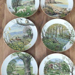 Op de afbeelding: Set van zes decoratieve borden met natuurtaferelen. Elk bord toont een ander landschap met bomen, bloemen en dieren. De borden zijn wit met een gouden rand.