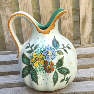 Op de afbeelding: Een witte keramische kan met een groen en bruin bloemenontwerp. De kan heeft een rond lichaam en een gebogen handvat.