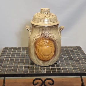 Op de afbeelding: Een bruin keramieken pot met deksel, met een verhoogd pompoenontwerp en het woord "RUMTOPS". De pot heeft een geribbelde textuur en kleine handvatten. Het deksel heeft een decoratief bloemmotief.