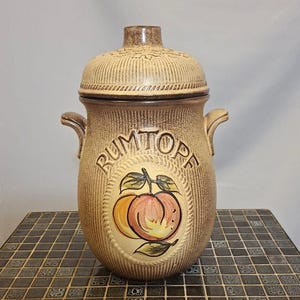 May include: A vintage ceramic jar with a lid, featuring a textured brown exterior and a decorative oval with the word "RUMTOPE" and a painted peach design. The jar has two small handles.