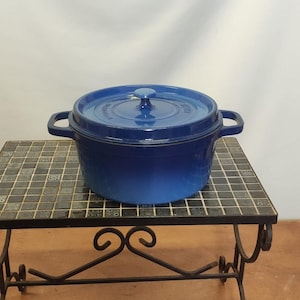 Op de afbeelding: Een blauwe geëmailleerde gietijzeren stoofpot met een deksel en twee handvatten. De pot staat op een zwarte metalen standaard met een betegelde bovenkant.