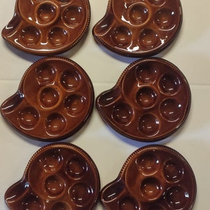 May include: Six brown ceramic escargot plates. Each plate has a snail shell shape with six circular indentations for holding snails. The plates have a glossy finish and a decorative edge.