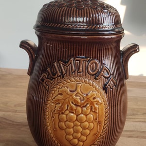 May include: Brown ceramic Rumtopf pot with a lid and handles. The pot features a textured surface with vertical lines and a circular emblem with grapes and the word "RUMTOPF".