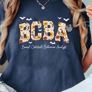 Op de afbeelding: Marineblauwe t-shirt met de tekst "BCBA" in een bloemenontwerp, met vleermuizen en de woorden "Board Certified Behavior Analyst" eronder. Een comfortabele, casual top.