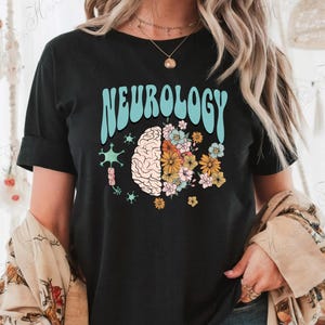 May include: Black t-shirt with the word "NEUROLOGY" in light blue, above a brain graphic with flowers. The design includes a brain, flowers, and neuron illustrations, creating a unique medical and floral theme.