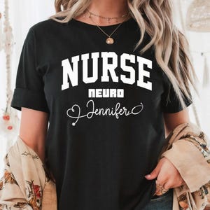 May include: Black t-shirt with the word "NURSE" in large white block letters. Below it, the word "NEURO" is centered, and the name "Jennifer" is written in cursive with a stethoscope graphic. The shirt has rolled-up sleeves.