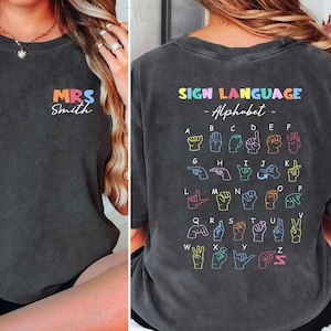以下が含まれることがあります： 前面にカラフルな文字で「MRS Smith」と書かれた濃い灰色のTシャツ。背中には、手話アルファベットが虹色の配色で表示され、「SIGN LANGUAGE - Alphabet -」の文字が手のジェスチャーの上に書かれています。