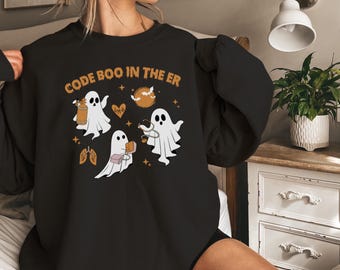 Pode incluir: Camisola preta com o texto "CODE BOO IN THE ER" em laranja. O design apresenta fantasmas de desenhos animados com equipamentos médicos, um coração, pulmões e uma lua. Um design divertido com tema de Halloween.