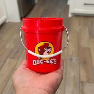 May include: A small, red bucket with a silver handle and a white grip. The bucket features a circular logo with a cartoon beaver and the text "BUC-EE'S" in white letters. The bucket is held in a person's hand.