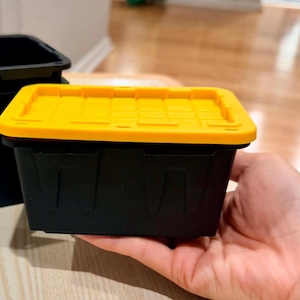 May include: A small rectangular storage container with a matte black base and a bright yellow snap-on lid. The lid features a grid-like embossed pattern and a slightly raised edge. The container has vertical structural ribs along its sides.
