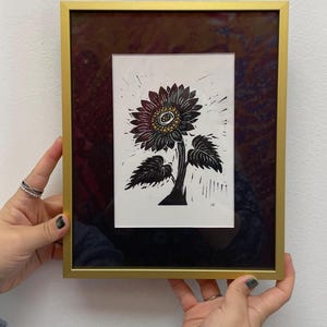 Framed black and gold sunflower linocut print with metallic center details, displayed in a modern gold frame against a white wall by someone holding the 8x10 inch frame to show size reference.