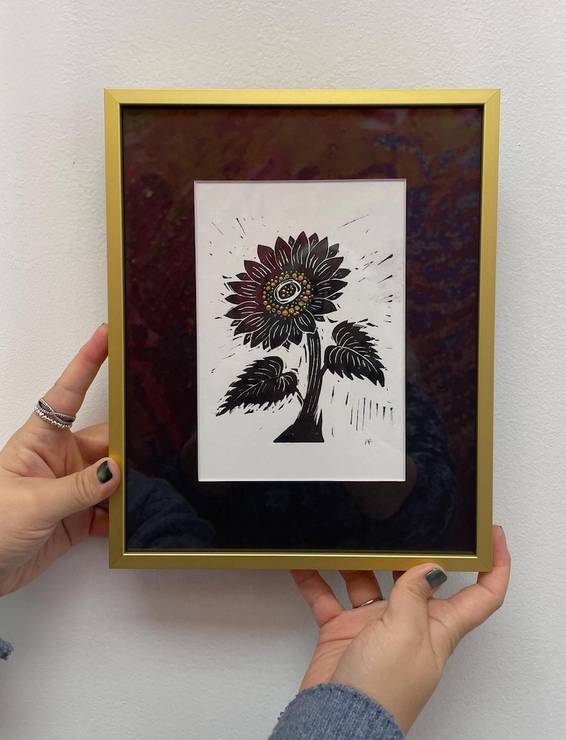 Framed black and gold sunflower linocut print with metallic center details, displayed in a modern gold frame against a white wall by someone holding the 8x10 inch frame to show size reference.