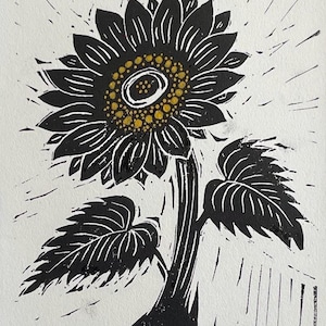 Black and white linocut print of a sunflower. The sunflower has a dark center with yellow dots, surrounded by black petals. The stem and leaves are also black, set against a white background. The artwork is signed in the bottom right corner.