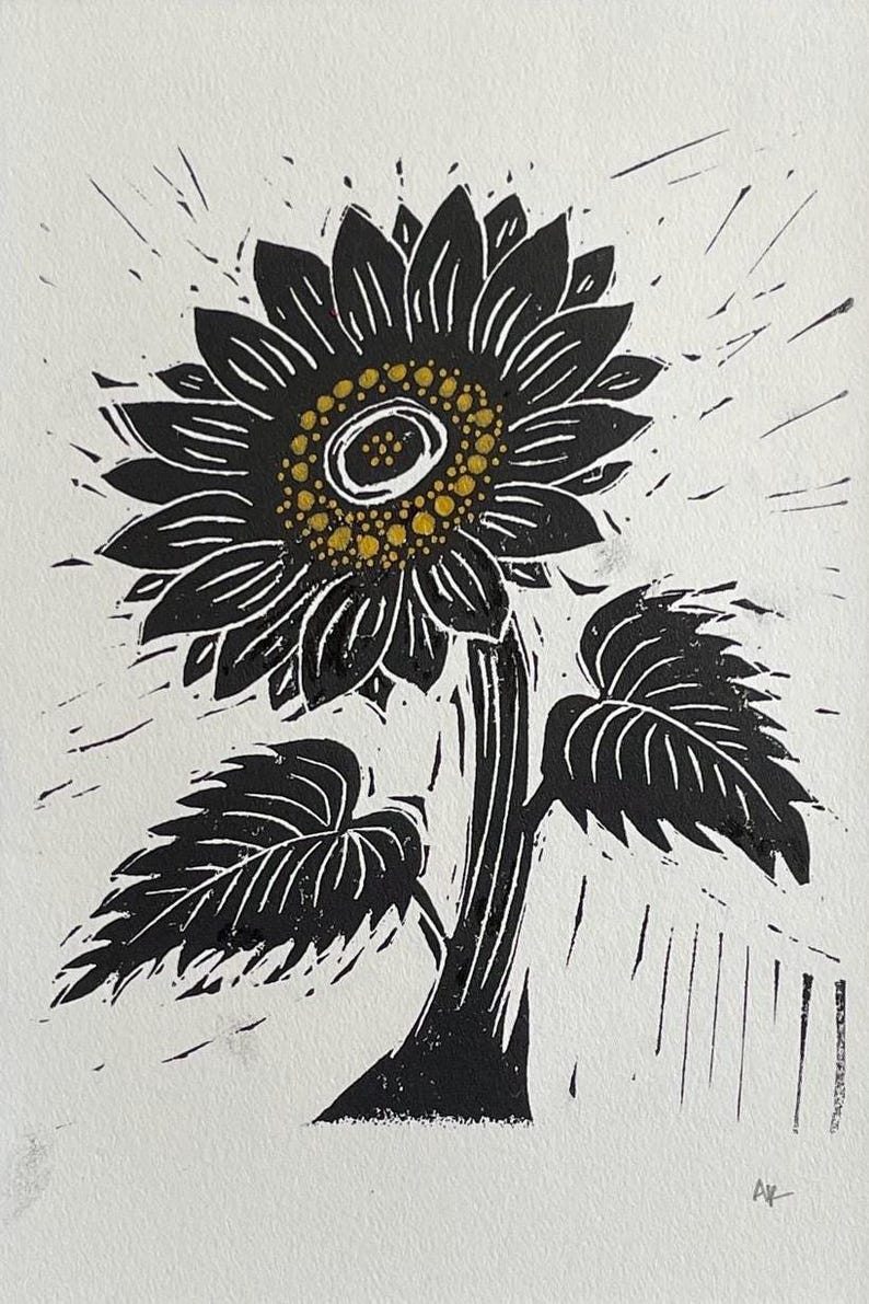 Black and white linocut print of a sunflower. The sunflower has a dark center with yellow dots, surrounded by black petals. The stem and leaves are also black, set against a white background. The artwork is signed in the bottom right corner.