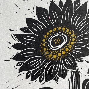 Black and white linocut print of a sunflower. The sunflower's petals are black with white lines, and the center is yellow. The print is on white paper, with black ink splatters around the flower.