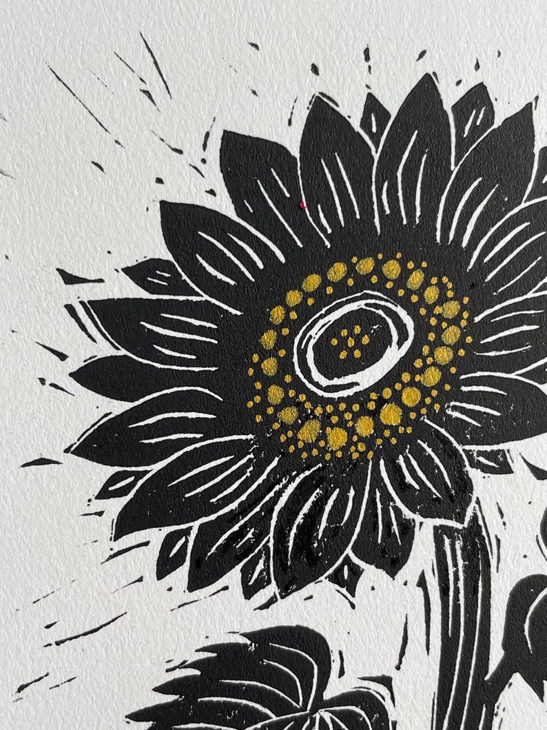Black and white linocut print of a sunflower. The sunflower's petals are black with white lines, and the center is yellow. The print is on white paper, with black ink splatters around the flower.