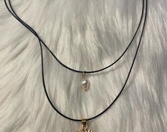 Puede incluir: Un collar de dos capas con un cordón negro y dos colgantes. El colgante inferior es un amuleto de ojo turco dorado con un centro azul. El colgante superior es una perla blanca.
