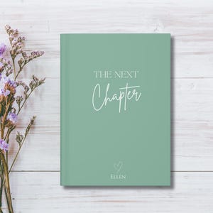 May include: A sage green journal with the words "THE NEXT Chapter" in white script. Below the text is a small heart and the name "ELLEN". The journal is on a white wooden surface with purple flowers.
