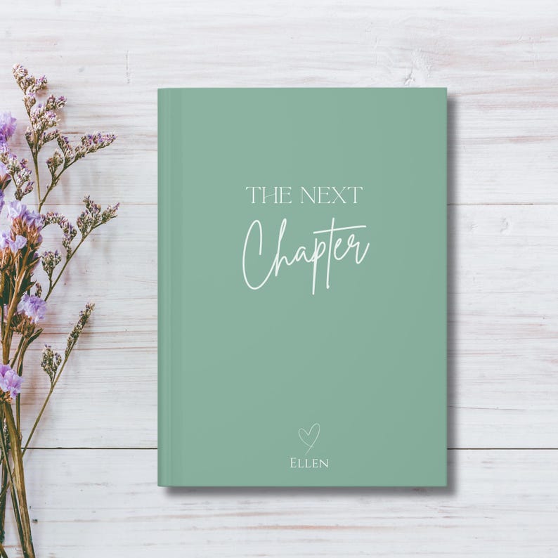 May include: A sage green journal with the words "THE NEXT Chapter" in white script. Below the text is a small heart and the name "ELLEN". The journal is on a white wooden surface with purple flowers.