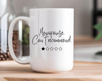 May include: White ceramic mug with the text "Menopause. Can't recommend." in black script. Below the text is a rating of one black star and four empty stars. The mug has a large handle.
