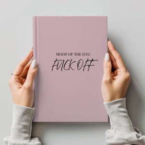 May include: A dusty rose hardcover journal featuring the black printed text MOOD OF THE DAY: FUCK OFF centered on the front cover. The book has a smooth, matte finish and a vertical spine on the left side, presented in a clean, minimalist style.