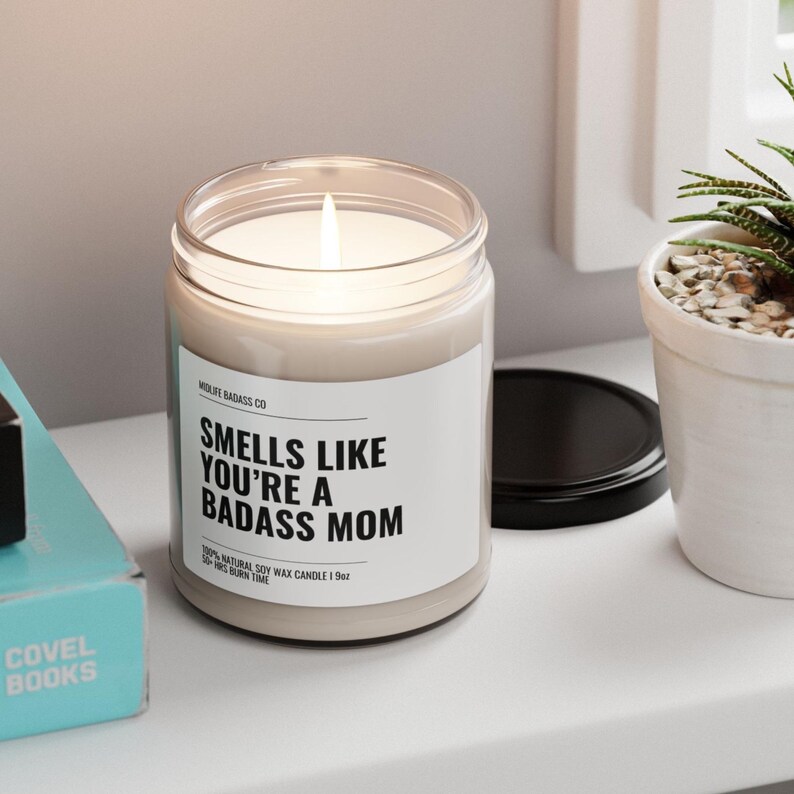 May include: A clear glass candle jar with a white label that reads "Smells Like You're A Badass Mom". The candle is lit and the jar is sitting on a white surface.