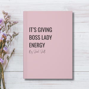Puede incluir: Un cuaderno rosa con el texto "IT'S GIVING BOSS LADY ENERGY" en negro. El nombre del autor, Sarah Smith, está escrito debajo. El cuaderno está sobre una superficie de madera blanca con flores moradas en el fondo.
