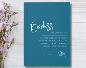 May include: A teal notebook with the word "Badass" in cursive script. The definition of "Badass" is printed below, along with the name "Stacey". The notebook is on a white wooden surface with purple flowers.