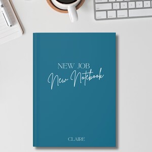 May include: A teal notebook with the words "NEW JOB" and "New Notebook" in white script. The name "CLAIRE" is printed at the bottom. The notebook is on a white surface, with a keyboard and a coffee cup in the background.