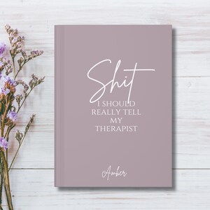 May include: A dusty mauve hardcover journal featuring the white text "Shit I SHOULD REALLY TELL MY THERAPIST" in a mix of script and serif fonts, with the name "Amber" printed in a script font near the bottom center of the front cover.