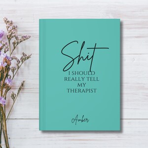 May include: A teal hardcover journal with black text that reads "Shit I SHOULD REALLY TELL MY THERAPIST" in a mix of script and serif fonts, with the name "Amber" printed in script at the bottom center of the front cover.