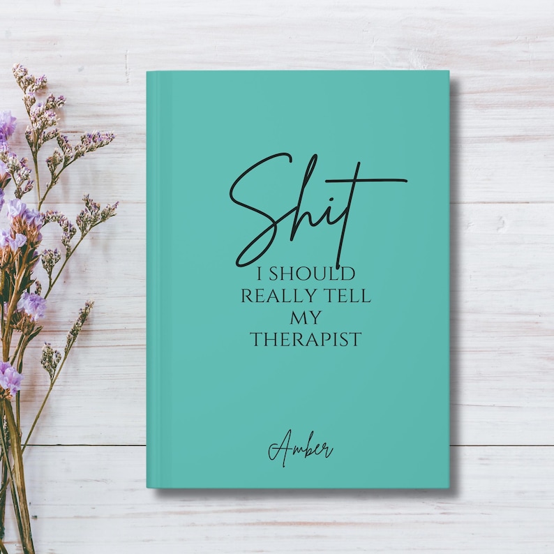 May include: A teal hardcover journal with black text that reads "Shit I SHOULD REALLY TELL MY THERAPIST" in a mix of script and serif fonts, with the name "Amber" printed in script at the bottom center of the front cover.