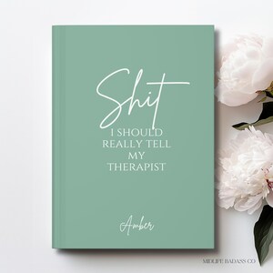 May include: A sage green hardcover journal featuring the white text "Shit I SHOULD REALLY TELL MY THERAPIST" in a mix of script and sans-serif fonts, with the name "Amber" printed below and white peony flowers visible on the right side.