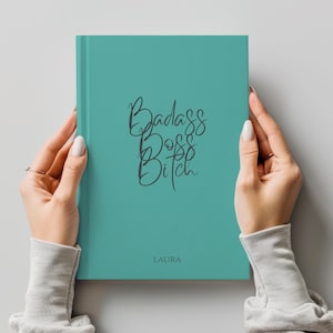 May include: A teal journal with the words "Badass Boss Bitch" in cursive script. The name "Laura" is printed below the text. The journal is held by a person.