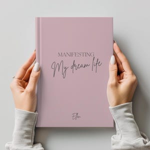 May include: A pink journal with the words "MANIFESTING My dream life" in dark gray script. The journal is held by two hands with long, manicured nails. The name "Ellen" is printed at the bottom.