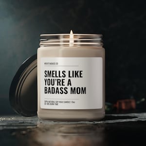 May include: A beige candle with a black label that reads "Smells Like You're A Badass Mom". The candle is 100% natural soy wax and has a 50-hour burn time.