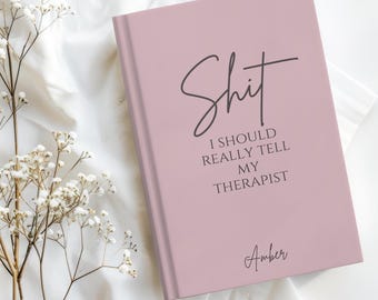 May include: A dusty rose hardcover journal featuring the black printed text "Shit I SHOULD REALLY TELL MY THERAPIST" and the name "Amber" in a script font at the bottom. The notebook is positioned next to a sprig of small white flowers.