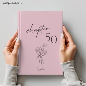May include: A light pink hardcover book featuring the black text "chapter 50" and the name "Sophie" in a cursive font. Below the text is a black line drawing of a small bouquet of three flowers. The brand name "midlife badass co" is at the top.