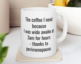 May include: White ceramic coffee mug with the text "The coffee I need because I was wide awake at 3am for hours - thanks to perimenopause." The mug is on a beige coaster.