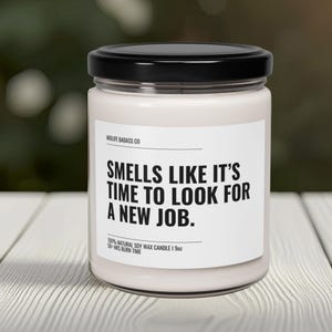 Pode incluir: Uma vela de cera de soja branca num frasco de vidro com tampa preta. O r&oacute;tulo diz "SMELLS LIKE IT'S TIME TO LOOK FOR A NEW JOB." A vela tem 255 gramas e um tempo de queima de mais de 50 horas. A marca &eacute; Midlife Badass Co.