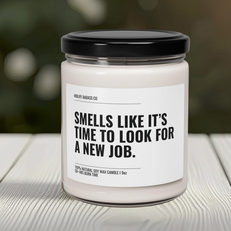 Pode incluir: Uma vela de cera de soja branca num frasco de vidro com tampa preta. O r&oacute;tulo diz "SMELLS LIKE IT'S TIME TO LOOK FOR A NEW JOB." A vela tem 255 gramas e um tempo de queima de mais de 50 horas. A marca &eacute; Midlife Badass Co.