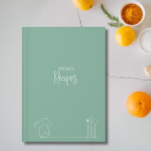 Puede incluir: Un libro de recetas turquesa con el texto "MOM'S Recipes" en cursiva. Un gorro de chef, un tenedor, un cuchillo y una cuchara están dibujados en blanco en la parte inferior. Limones, naranjas y especias están dispuestas alrededor del libro.