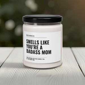 May include: A white soy wax candle with a black lid and label that reads "Smells Like You're A Badass Mom". The candle is 19 ounces and has a 50-hour burn time.