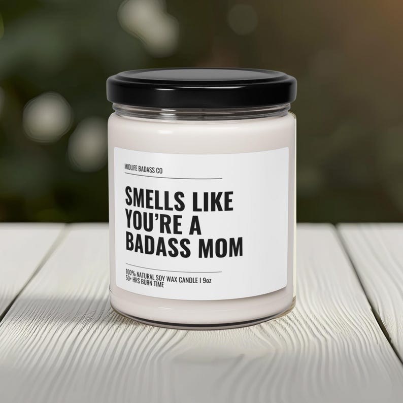 May include: A white soy wax candle with a black lid and label that reads "Smells Like You're A Badass Mom". The candle is 19 ounces and has a 50-hour burn time.