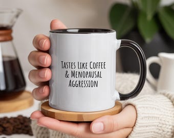 May include: White coffee mug with a black interior and handle, resting on a wooden coaster. The mug features the text "Tastes Like Coffee & Menopausal Aggression" in black lettering. The mug is held by a person.