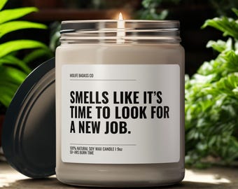 Op de afbeelding: Een brandende sojawas kaars in een glazen pot met een zwart deksel. Het etiket luidt "SMELLS LIKE IT'S TIME TO LOOK FOR A NEW JOB." De kaars is 255g en heeft een brandtijd van 50 uur.
