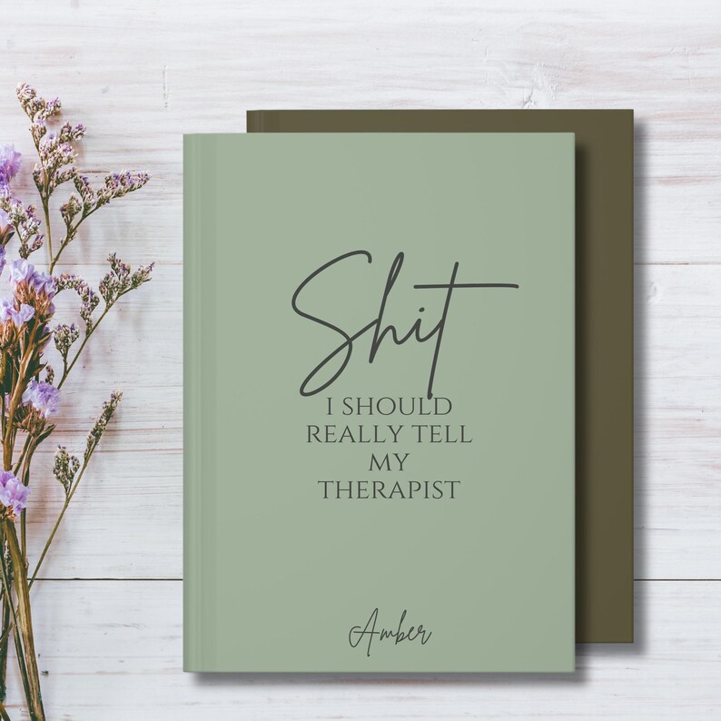 May include: A sage green hardcover journal with dark gray text that reads "Shit I SHOULD REALLY TELL MY THERAPIST" and the name "Amber" at the bottom. The journal is positioned in front of a dark olive green notebook, with dried purple flowers.